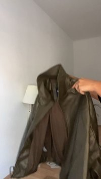 Giacca in pelle verde oliva con colletto alto, chiusura a zip e texture liscia. Fodera interna marrone. La giacca è sostenuta su uno sfondo chiaro.