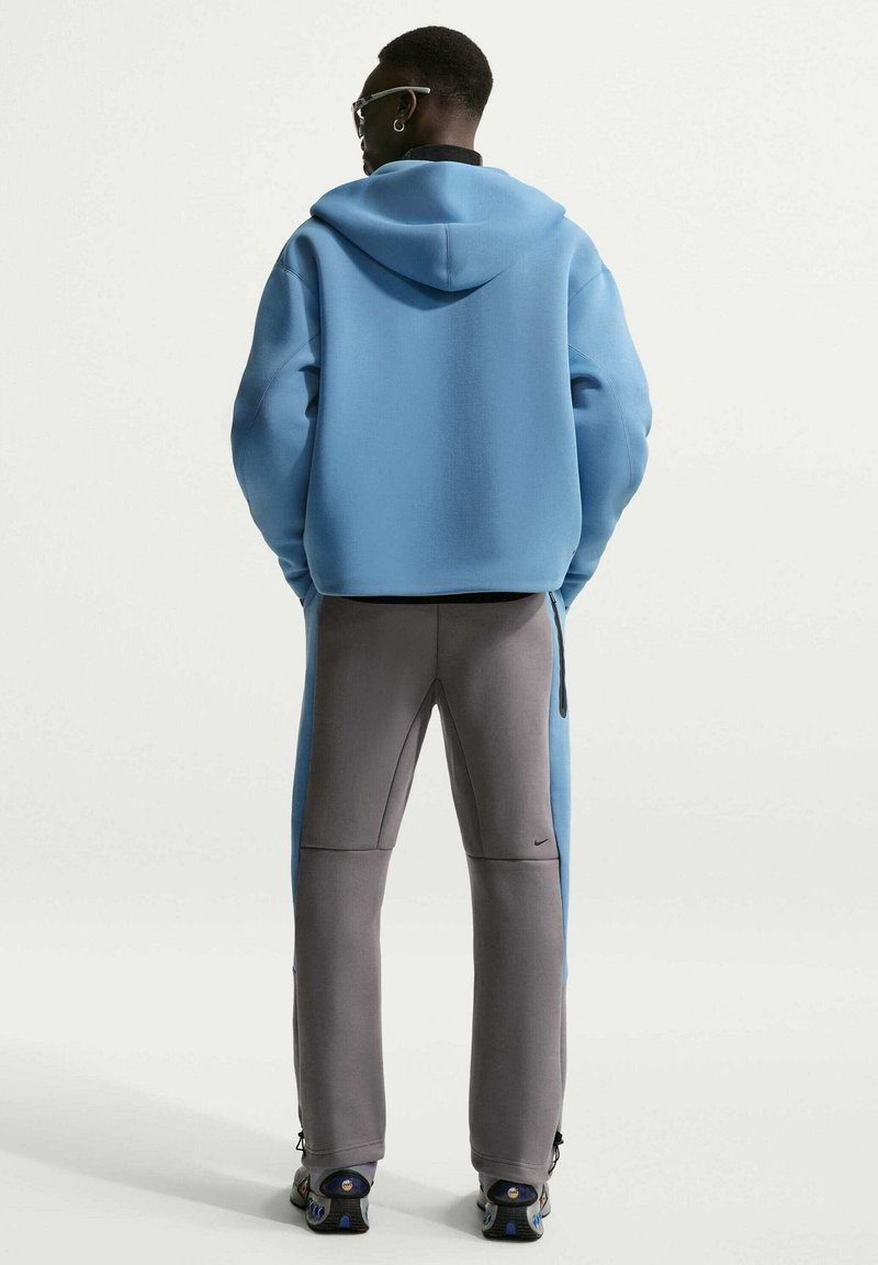 Mann trägt hellblauen Hoodie und graue Jogginghose mit blauen Seitenteilen, steht mit dem Rücken zur Kamera vor einfarbigem Hintergrund.