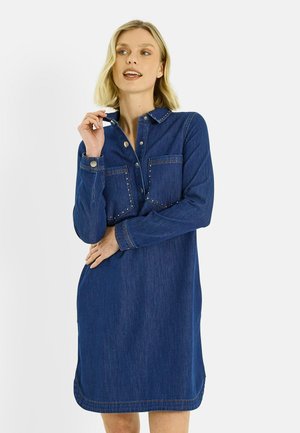 Femme portant une robe en denim à manches longues avec des boutons-pression et des poches poitrine, posant avec une main près du col sur un fond uni.