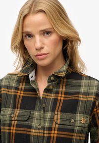 Plaid button-up shirt featuring green, black, and orange colors, with two chest pockets and a collar. Made of textured fabric, detailed stitching.
