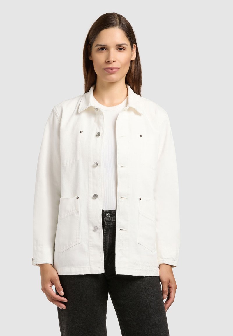 Femme aux cheveux bruns raides portant une veste blanche boutonnée, une chemise blanche et un jean noir, debout devant un fond gris clair.