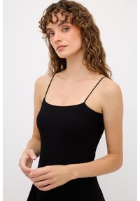 Abito nero aderente con sottili spalline, texture a coste e cuciture strutturate, indossato da una modella con capelli ricci su uno sfondo neutro.