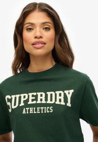 Camiseta de algodón verde con manga corta, cuello redondo, que presenta un gran texto blanco "SUPERDRY ATHLETICS" en el pecho.