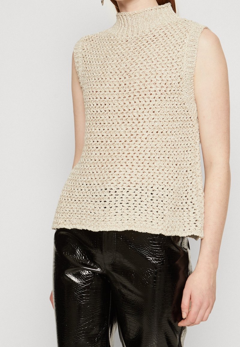 Haut sans manches beige en maille avec col montant associé à un pantalon noir brillant en similicuir à texture croco.