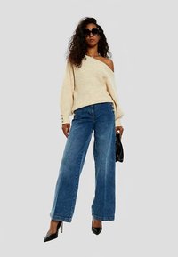 Kreminės spalvos perdidintas megztinis, viena petį atidengiantis, derinamas su plačiomis, aukšto liemens mėlynomis džinsais, juodais aukštakulniais ir juoda rankine.