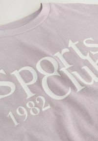 Lilla T-shirt med korte ærmer lavet af bomuld, prydet med fed hvid tekst, der står "Sports Club 1982" i en moderne skrifttype.
