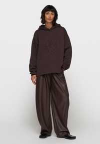Bluza z kapturem w ciemnym brązie z wytłoczonym logo i luźnym krojem, zestawiona z szerokimi spodniami z brązowej sztucznej skóry i czarnymi butami typu slip-on.