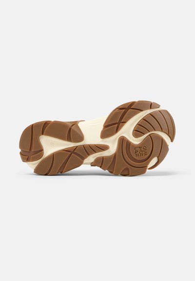 Zool van een schoen met bruin rubberen profiel en crème tussenzool, voorzien van gebogen groeven en een cirkelvormig logodetail op het voorvoetgedeelte.