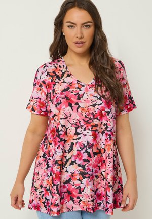 Frau mit langem braunem Haar, die ein lockeres, kurzärmeliges V-Ausschnitt-Oberteil mit rosa und roten Blumenmustern trägt und vor einem schlichten hellen Hintergrund steht.