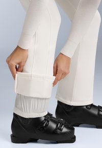 Bocanci de schi negri cu trei catarame ajustabile, asortați cu pantaloni de culoare crem cu manșete elastice și o textură netedă.