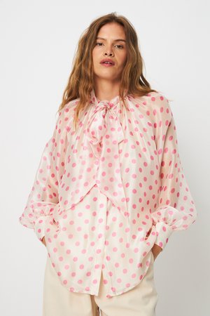 Kvinde iført gennemsigtig hvid bluse med lyserøde prikker og stor sløjfe ved halsen, lange puffede ærmer, hænder i beige bukselommer.