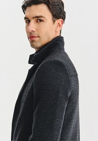 Czarny teksturowany płaszcz z podniesionym kołnierzem i subtelnym wzorem, charakteryzujący się dopasowanym krojem i gładkim materiałem. Dopasowana sylwetka pokazana z boku.