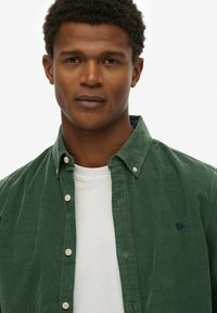 Groene corduroy shirt met een button-down kraag, voorzien van witte knopen en een marineblauw geborduurd logo op de borst, laag over een wit t-shirt.