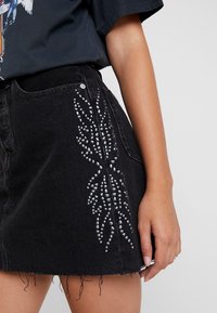 Detailní záběr na osobu v černé džínsové sukni se stříbrnými korálkovými vzory listů a roztrhaným lemem, v kombinaci s tmavým tričkem s grafikou.