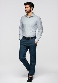 Homme debout, les mains dans les poches, portant une chemise à boutons gris clair, un jean bleu foncé, une ceinture noire et des mocassins noirs sur un fond uni.