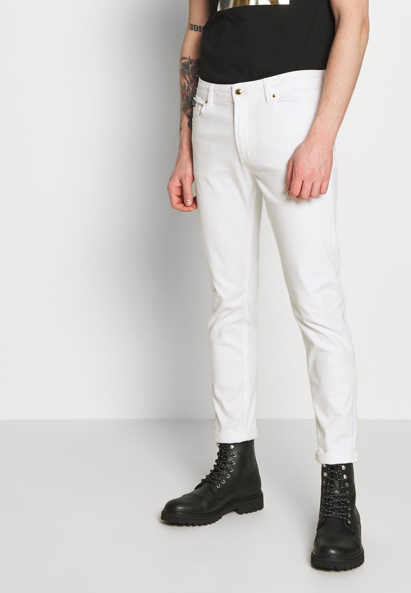 Personne portant un jean blanc slim ajusté avec des revers aux chevilles, des bottes en cuir noires à lacets et une chemise noire, avec un tatouage visible sur le bras.