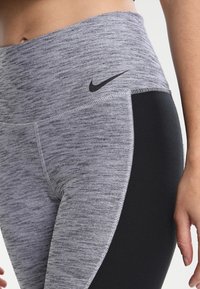 Szare i czarne legginsy sportowe z wysokim stanem, teksturowana tkanina oraz małe czarne logo Nike po lewej stronie u góry.