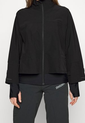 Person trägt eine schwarze Jacke mit Reißverschluss und verlängerten Ärmeln sowie schwarze Hose mit "zimtstern"-Logo am linken Oberschenkel.