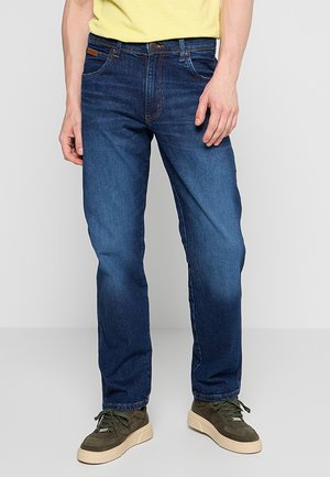 Jeans da uomo in denim blu scuro con vestibilità dritta, caratterizzati da cuciture a contrasto, passanti per cintura e cinque tasche. Indossati con sneakers verdi.