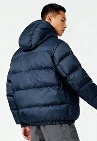 Giacca imbottita blu navy con cappuccio, caratterizzata da sezioni trapuntate e orlo elasticizzato. Tessuto liscio, design leggero, adatta per il freddo.