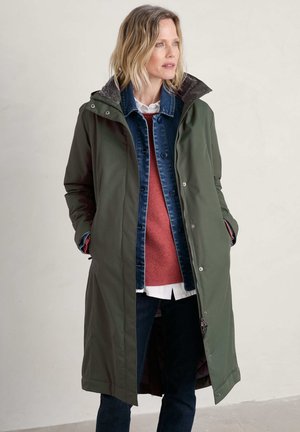 Long, olive-green waterproof coat with a zip front, silver buttons, and a textured interior. Worn over a denim jacket and red sweater.