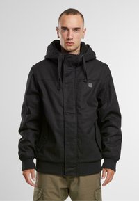 Chaqueta negra con capucha, cierre frontal, puños elásticos y un acabado texturizado. Presenta un parche con el logo y bolsillos laterales para mayor funcionalidad.