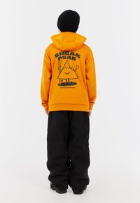 Jasne oranžová mikina s grafikou hory a postavy na chrbte, kombinovaná s čiernymi voľnými nohavicami a čiernou čiapkou.
