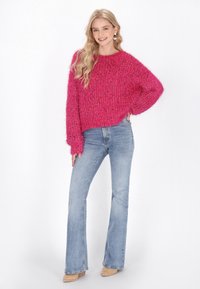 Magenta fuzzy gebreide trui met een ontspannen pasvorm en verlaagde schouders, gecombineerd met lichtblauwe flared jeans en beige enkellaarsjes.