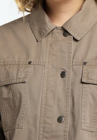 Khakifarbene Baumwoll-Hemdenjacke mit einem zuknöpfbaren Kragen, zwei Brusttaschen und silberfarbenen Metallakzenten. Weicher, strukturierter Stoff.