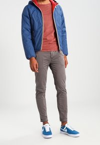 Blå pufferjacka med röd insida, grå långärmad tröja, ljusgrå byxor och blå sneakers med vita accenter och snörning.