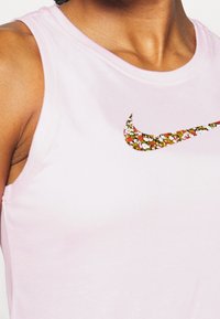 Jasnoróżowa bezramienna koszulka sportowa z teksturowanym, kwiatowym logo Nike na klatce piersiowej. Miękki, elastyczny materiał o gładkim wykończeniu.