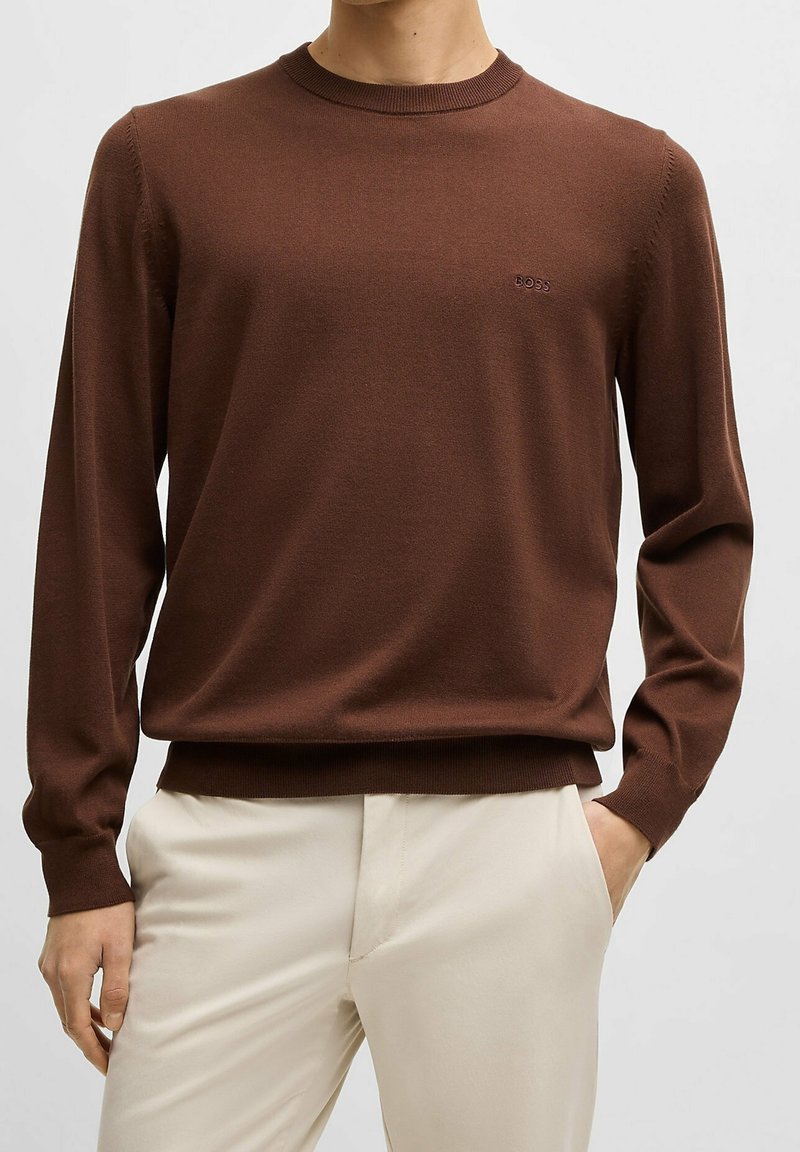 Homme portant un pull marron à manches longues et col rond avec le logo "BOSS" sur la poitrine et un pantalon beige clair, main dans la poche, debout devant un fond uni.