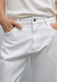 Le jean en denim blanc présente un design classique avec des poches avant et une fermeture à bouton métallique. Le tissu a une surface lisse et texturée.