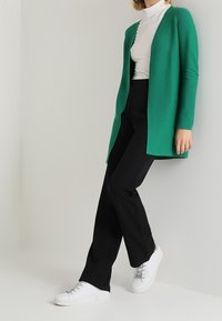 Grüne Strickjacke, lange Ärmel, offene Vorderseite, getragen über einem weißen Rollkragenpullover. Schwarze Hose und weiße Turnschuhe vervollständigen das Outfit. Glatte Textur.