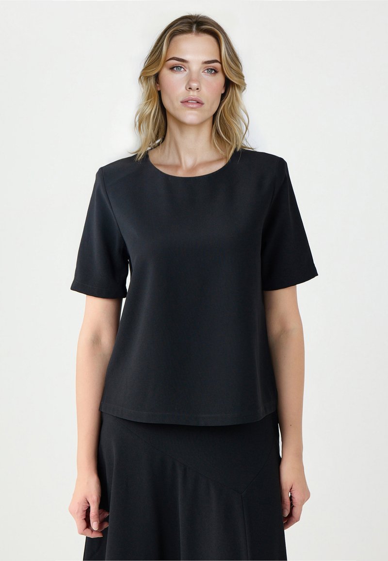 Kobieta o blond włosach, ubrana w prostą czarną bluzkę z krótkim rękawem i czarną spódnicę, na jasnym tle, zwrócona przodem, z neutralnym wyrazem twarzy.