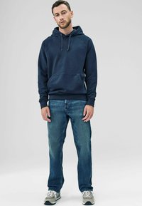 Felpa blu navy con tasca a marsupio, polsini a coste e cappuccio con cordini, abbinata a jeans azzurri e scarpe da ginnastica grigie.
