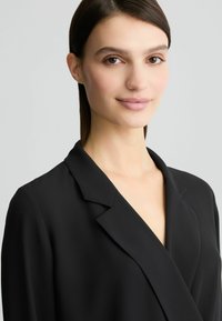 Čierny blazer s klopou, hladkou textúrou a priliehavým strihom, ktorý zvýrazňuje výstrih s hlbokým V-krkom.