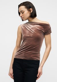 Femme aux cheveux courts et foncés portant un haut marron en velours à épaules dénudées et un pantalon noir, regardant vers le bas à sa gauche sur un fond uni.