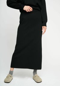Svart stickad midikjol med ett figurnära midja, slät textur och golvlång design. Parat med grå strumpor och beige slip-on skor.