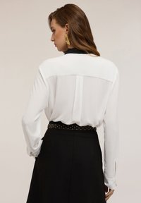 Blusa bianca con colletto nero, maniche lunghe con fessure, abbinata a una gonna nera e una cintura con borchie. Materiale morbido e design pulito.