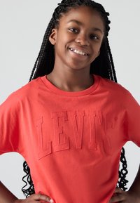 Camiseta de algodón roja con cuello redondo y mangas cortas, que presenta el texto en relieve "LEVI'S" en el frente. Textura suave y ajuste casual.