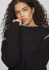 Čierny pletený sveter s dlhými rukávmi, s rebrovanou textúrou a bielym prešitím pozdĺž výstrihu a ramenných švov.