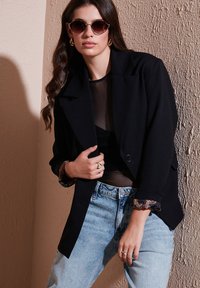 Černý oversized blejzr s klopovým límcem a knoflíkovým zapínáním, kombinovaný se světle modrými jeansy s vysokým pasem. Pod ním je průhledný černý top s vzorovanými manžetami.