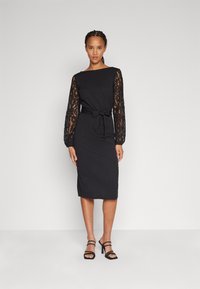 Robe noire avec des manches longues en dentelle, silhouette ajustée, ourlet droit et une ceinture à nouer à la taille. Portée avec des talons noirs à lanières.