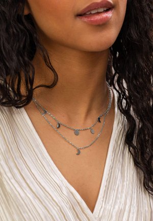 Collier en argent superposé avec des pendentifs en forme de cercle et de croissant. Les chaînes ont une finition lisse, contrastant avec un haut texturé de couleur claire.