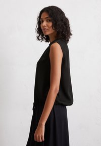 Vrouw met krullend donker haar, gekleed in een mouwloze zwarte top en rok, staat zijwaarts tegen een effen witte muur.