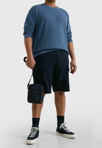 Blau strukturierter Pullover, marineblaue Shorts, trägt eine kleine marineblaue Tasche. Weiße Akzente an Schuhen und Socken. Lässiges Outfit mit einem entspannten Schnitt.