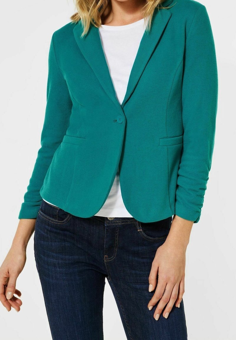 Teal kavaj med en figursydd design, enkel knappstängning, slagkrage och två framfickor. Bärs över en vit skjorta och mörka jeans.