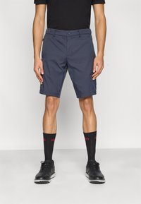 Temno modre kratke hlače s kratkim krojem, z zaponko na gumb, zankami za pas in stranskimi žepi, kombinirane s črnimi čevlji in nogavicami do kolen.