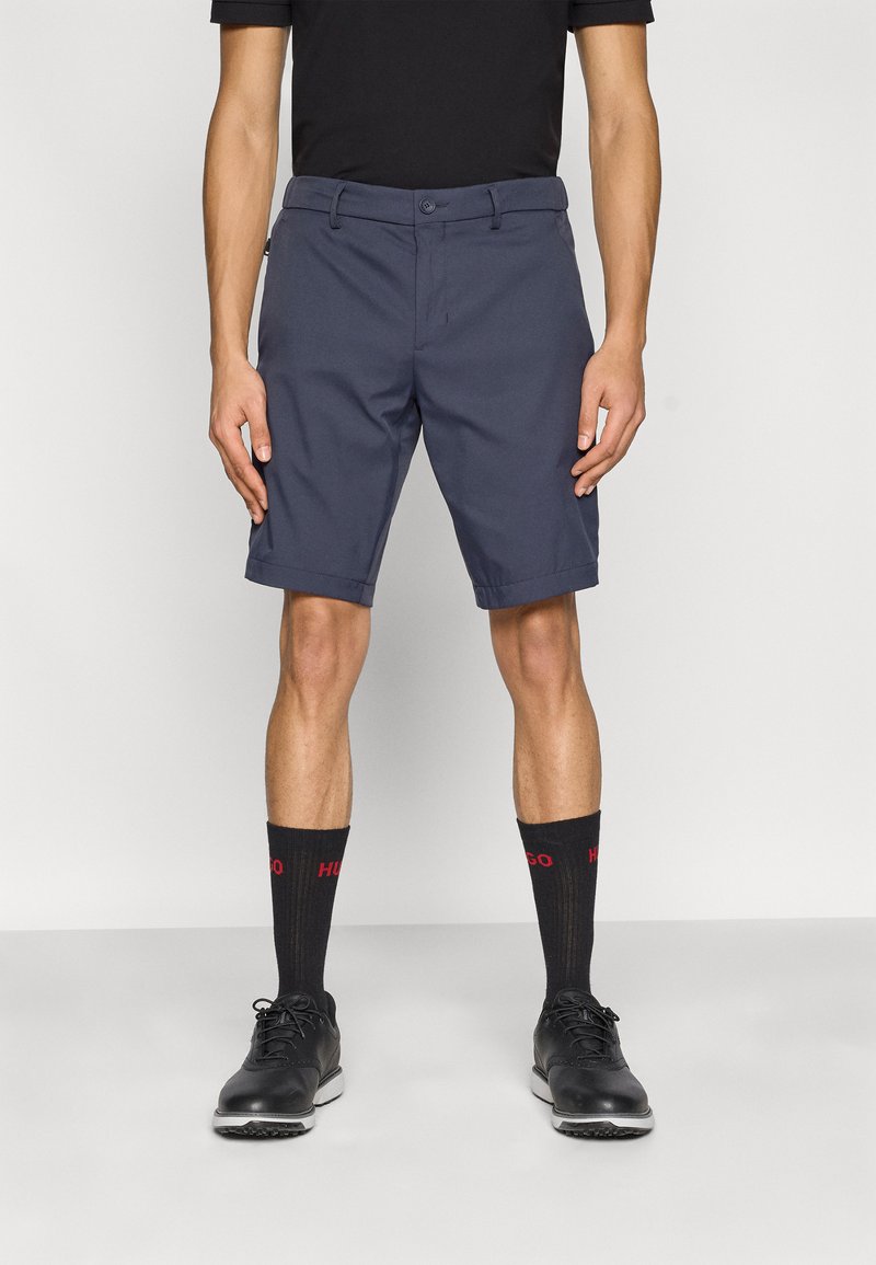 Temno modre kratke hlače s kratkim krojem, z zaponko na gumb, zankami za pas in stranskimi žepi, kombinirane s črnimi čevlji in nogavicami do kolen.